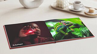 Perspektivische Aufnahme eines aufgeschlagenen, quadratischen Premium-Fotobuchs auf einem beigen Tisch. Die linke Seite zeigt eine Frau, die einen rot leuchtenden Rubin vor ihr Gesicht hält, und trägt den Text 'RubyPaper'. Die rechte Seite zeigt ein Nahaufnahme-Foto eines grünen Chamäleons im Blätterwald. Im Hintergrund sind eine ovale Skulptur, eine moderne Kerze und ein Kaffeetablett mit Makrone zu sehen.