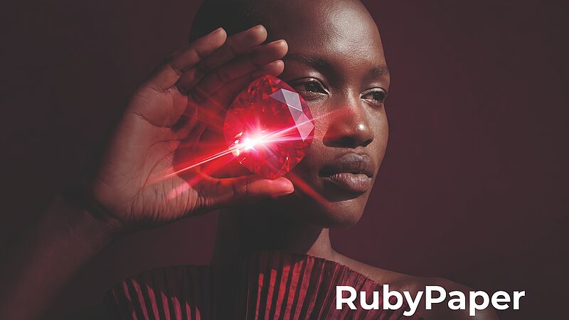 Nahaufnahme einer Person vor dunklem Hintergrund, die einen großen, facettierten Rubin vor das rechte Auge hält. Der rote Edelstein reflektiert ein helles Licht mit strahlenförmigen Lichteffekten. Die Person trägt ein gefaltetes Kleidungsstück in Burgunderrot